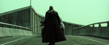 Movie still from “The Matrix Reloaded” (2003), directed by Lana Wachowski – A man holding a baseball bat while standing next to a car; Wide shot, Low angle