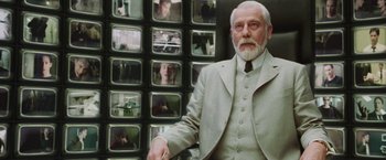 Movie still from “The Matrix Reloaded” (2003), directed by Lana Wachowski – An older man in a suit and tie in front of a wall of pictures; Medium shot, Over the shoulder angle