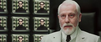 Movie still from “The Matrix Reloaded” (2003), directed by Lana Wachowski – An older man in a suit and tie in front of a picture of a man; Close Up shot, Over the shoulder angle