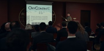 Movie still from “Dopesick” (2021), directed by Danny Strong – A man standing in front of a group of people in front of a projection screen; Extreme Wide shot, Over the shoulder angle