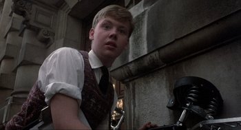 Movie still from “The Meaning of Life” (1983), directed by Terry Jones – A young man in a vest and tie standing in front of a building; Medium shot, Low angle