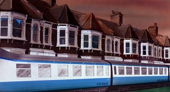 Movie still from “The Meaning of Life” (1983), directed by Terry Jones – A blue and white train traveling down train tracks next to houses; Extreme Wide shot, Low angle