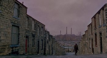 Movie still from “The Meaning of Life” (1983), directed by Terry Jones – A person walking down a street near a bunch of buildings; Extreme Wide shot, High angle