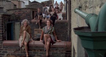 Movie still from “The Meaning of Life” (1983), directed by Terry Jones – A group of people sitting on the side of a brick wall; Wide shot, High angle