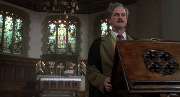 Movie still from “The Meaning of Life” (1983), directed by Terry Jones – An old man in a suit and tie in a church with stained glass windows; Medium shot, Low angle