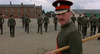 Movie still from “The Meaning of Life” (1983), directed by Terry Jones – A man in a uniform holding a baseball bat in front of a group of soldiers; Medium shot, Low angle