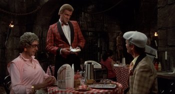 Movie still from “The Meaning of Life” (1983), directed by Terry Jones – A man in a red and white checkered jacket standing at a table; Medium shot, Over the shoulder angle