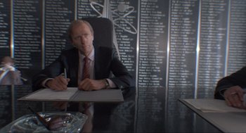 Movie still from “The Meaning of Life” (1983), directed by Terry Jones – A man sitting at a table in front of a wall of names; Medium shot, Over the shoulder angle