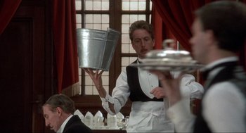 Movie still from “The Meaning of Life” (1983), directed by Terry Jones – A man holding a metal bucket over his head in a restaurant; Medium shot, Low angle