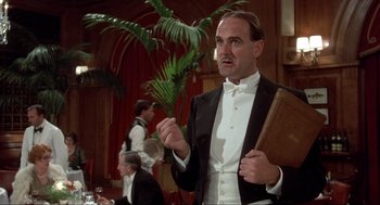 Movie still from “The Meaning of Life” (1983), directed by Terry Jones – A man in a tuxedo holding a book; Medium shot, Low angle