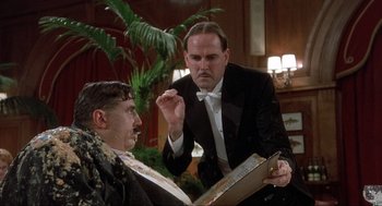 Movie still from “The Meaning of Life” (1983), directed by Terry Jones – A man in a suit and bow tie is looking at a book; Medium shot, Low angle