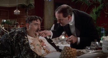 Movie still from “The Meaning of Life” (1983), directed by Terry Jones – A man in a suit and tie is feeding a man a piece of cake; Medium shot, Over the shoulder angle