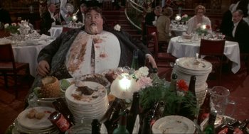 Movie still from “The Meaning of Life” (1983), directed by Terry Jones – A man sitting at a table with a lot of food on it; Medium shot, High angle