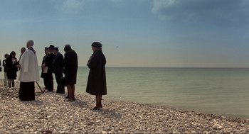 Movie still from “The Meaning of Life” (1983), directed by Terry Jones – A group of people standing on top of a sandy beach; Extreme Wide shot, High angle