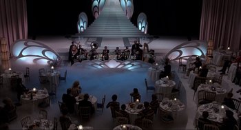 Movie still from “The Meaning of Life” (1983), directed by Terry Jones – A group of people sitting at tables in front of a staircase; Extreme Wide shot, High angle