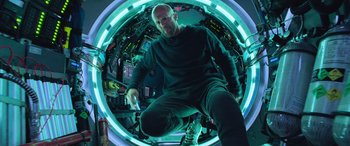 Movie still from “The Meg” (2018), directed by Jon Turteltaub – A bald man in a black shirt is in a room with lights; Medium shot, Low angle