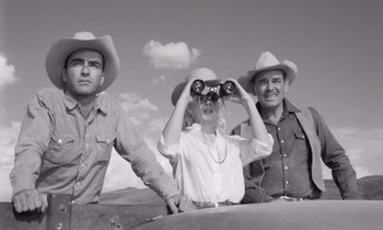 Movie still from “The Misfits” (1961), directed by John Huston – A woman looking through binoculars while standing next to two men in cowboy hats; Medium shot, Low angle