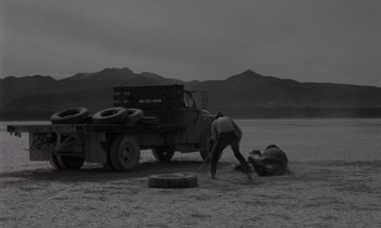 Movie still from “The Misfits” (1961), directed by John Huston – A man in black pants and a white shirt is on the ground next to a truck; Wide shot, High angle