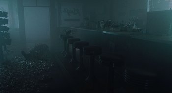 Movie still from “The Mist” (2007), directed by Frank Darabont – A dark room filled with black stools and a counter; Wide shot, High angle
