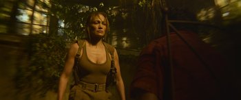 Movie still from “The Mother” (2023), directed by Niki Caro – A woman in a tan tank top and brown pants standing next to a tree; Medium shot, Over the shoulder angle