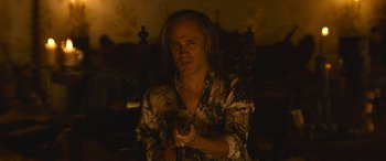 Movie still from “The Mother” (2023), directed by Niki Caro – A man sitting in a chair holding a remote control; Medium shot, Over the shoulder angle