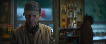Movie still from “The Mother” (2023), directed by Niki Caro – An older man wearing a hat and a jacket; Close Up shot, Over the shoulder angle