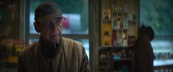 Movie still from “The Mother” (2023), directed by Niki Caro – An older man wearing a hat and a vest; Close Up shot, Over the shoulder angle