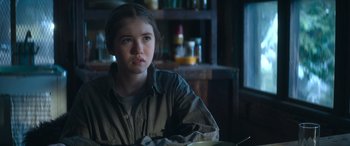 Movie still from “The Mother” (2023), directed by Niki Caro – A young girl sitting at a table in a room; Close Up shot, Over the shoulder angle