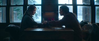 Movie still from “The Mother” (2023), directed by Niki Caro – Two people sitting at a table in front of a window; Medium shot, Over the shoulder angle