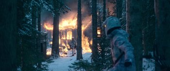 Movie still from “The Mother” (2023), directed by Niki Caro – A man in a helmet standing in front of a burning house in the woods; Wide shot, Over the shoulder angle