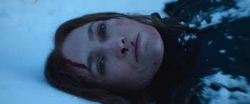Movie still from “The Mother” (2023), directed by Niki Caro – A young woman with a bloody nose laying in the snow; Close Up shot, Overhead angle