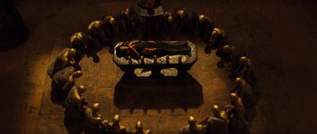 Movie still from “The Mummy” (1999), directed by Stephen Sommers – An image of a person laying on a bench in a circle of gold statues; Wide shot, Overhead angle