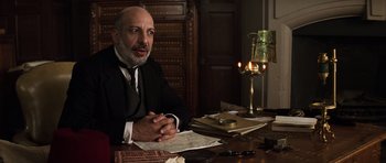Movie still from “The Mummy” (1999), directed by Stephen Sommers – An older man sitting at a table in front of papers; Medium shot, Low angle
