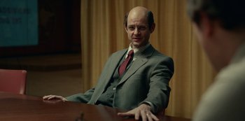 Movie still from “Dopesick” (2021), directed by Danny Strong – A man in a suit and tie sitting at a table; Medium shot, Over the shoulder angle