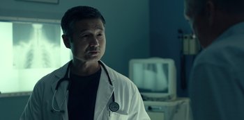 Movie still from “Dopesick” (2021), directed by Danny Strong – A man in a white lab coat stands in front of a television; Close Up shot, Low angle