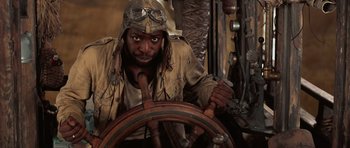 A man wearing a hat and a jacket sitting at the steering wheel of a boat
