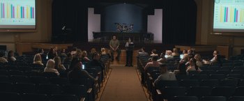 Movie still from “The Night House” (2020), directed by David Bruckner – A group of people sitting in a room with a stage; Extreme Wide shot, High angle