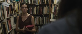 Movie still from “The Night House” (2020), directed by David Bruckner – A woman holding a book in front of bookshelves; Medium shot, Over the shoulder angle