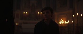 Movie still from “The Nun II” (2023), directed by Michael Chaves – A young man standing in the dark in front of candles; Medium shot, Over the shoulder angle