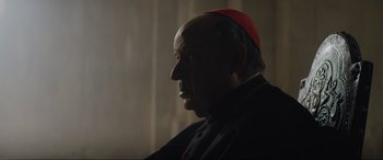 Movie still from “The Nun II” (2023), directed by Michael Chaves – An older man wearing a red hat and a black robe; Close Up shot, Over the shoulder angle