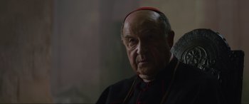 Movie still from “The Nun II” (2023), directed by Michael Chaves – An older man wearing a red hat and a black robe; Close Up shot, Over the shoulder angle