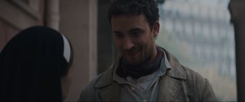 Movie still from “The Nun II” (2023), directed by Michael Chaves – A man with a scarf around his neck smiles for the camera; Close Up shot, Over the shoulder angle