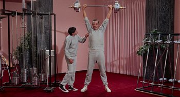 Movie still from “The Nutty Professor” (1963), directed by Jerry Lewis – A man lifts a barbell above his head while another man watches; Medium shot, Low angle