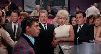 Movie still from “The Nutty Professor” (1963), directed by Jerry Lewis – A group of people sitting at a table in front of a crowd; Medium shot, Over the shoulder angle