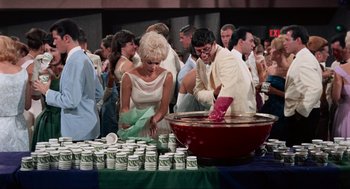 Movie still from “The Nutty Professor” (1963), directed by Jerry Lewis – A group of people standing around a table with cups on top of it; Wide shot, High angle