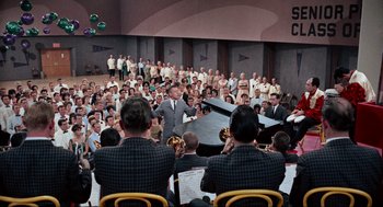 Movie still from “The Nutty Professor” (1963), directed by Jerry Lewis – A group of people sitting around a stage with a conductor; Wide shot, High angle