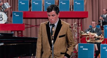 Movie still from “The Nutty Professor” (1963), directed by Jerry Lewis – A man in a suit and tie standing in front of a microphone; Medium shot, High angle