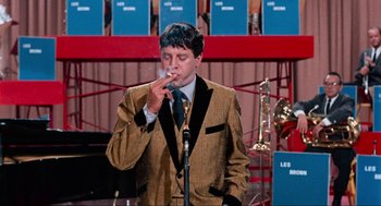 Movie still from “The Nutty Professor” (1963), directed by Jerry Lewis – A man smoking a cigarette while standing in front of microphones; Medium shot, Over the shoulder angle