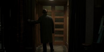 Movie still from “The Outfit” (2022), directed by Graham Moore – A man in a hat and a long coat standing in front of an open door; Wide shot, Over the shoulder angle