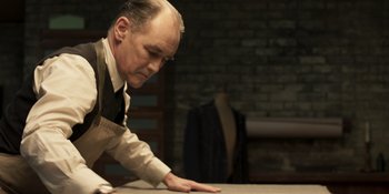 Movie still from “The Outfit” (2022), directed by Graham Moore – An older man leaning over a table in a room; Medium shot, High angle